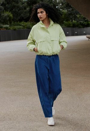 Femme aux cheveux bouclés marchant en extérieur, portant une veste vert clair, un pantalon bleu ample et des baskets blanches, les mains dans les poches.