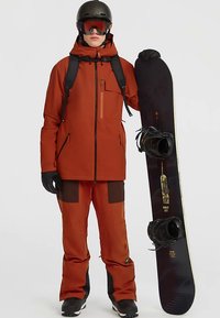 Persona in giacca da neve arancione, pantaloni, guanti neri, casco e occhiali da sci, che tiene una tavola da snowboard nera in piedi contro uno sfondo neutro.