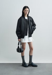 Cazadora negra tipo bomber con cremallera, vestido blanco, calcetas grises hasta la rodilla y zapatos negros. Accesorio: bolso pequeño negro con tachuelas.
