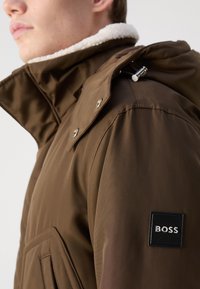 Smeđi šuškavni kaput s visokim ovratnikom koji sadrži bijelu ovčju podstavu. Ima bočne džepove i oznaku od tkanine s "BOSS" logotipom.