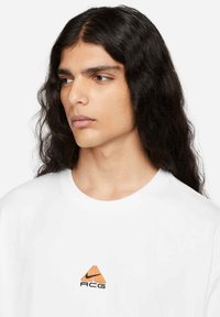 White cotton t-shirt featuring a small orange and black triangle logo with "ACG" text, round crew neckline, and short sleeves.