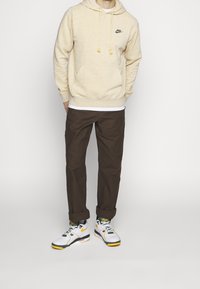Felpa beige maculata con cappuccio e lacci, pantaloni marroni e sneakers multicolori con dettagli bianchi, verdi e gialli.