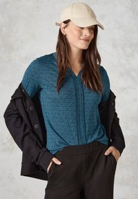 Helles blaues gemustertes Oberteil mit einem Reißverschluss vorne, kombiniert mit einer schwarzen Jacke und dunklen Hosen. Die Mütze ist beige und sorgt für einen lässigen Look.