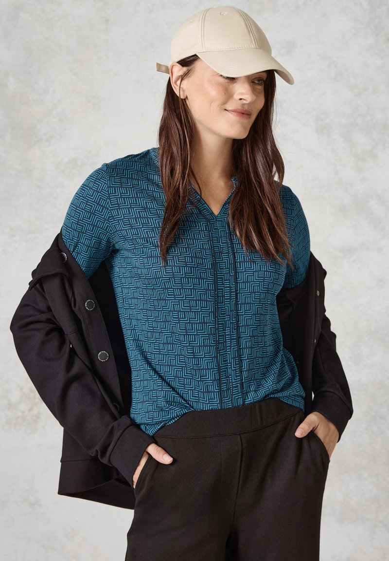Helles blaues gemustertes Oberteil mit einem Reißverschluss vorne, kombiniert mit einer schwarzen Jacke und dunklen Hosen. Die Mütze ist beige und sorgt für einen lässigen Look.