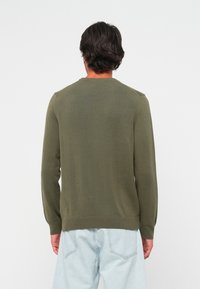 Man in een olijfgroen langemouwen trui en lichtblauwe jeans, staande met zijn rug naar de camera tegen een witte achtergrond.