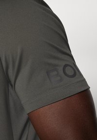 Pilkas sportinis marškinėliai su trumpomis rankovėmis, pasižymi tekstūruota medžiaga ir juodu "BOLD" logotipu ant rankovės, pabrėžiančiu sportišką dizainą.