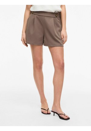 Femme portant un short plissé marron taille haute avec détail de boutons, associé à des sandales noires à petits talons, debout sur fond blanc.