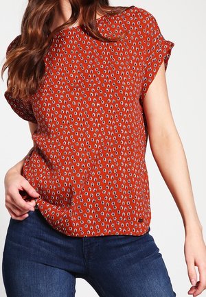 Femme portant un chemisier orange rouille à manches courtes avec de petits motifs de feuilles et un jean bleu foncé, debout devant un fond clair uni.