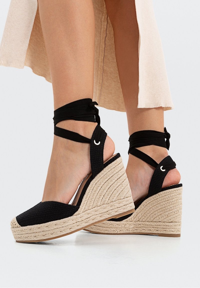 Sandales compensées en tissu noir tissé avec un semelle intermédiaire en corde naturelle, dotées de brides noires enroulées autour de la cheville et d'accents métalliques.