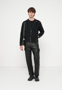 Svart jeansjacka med dragkedja och kontrasterande detalj på axeln, kombinerad med texturerade svarta läderbyxor och svarta sneakers. Minimalistisk design.