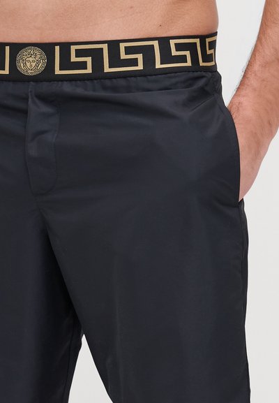 Shorts de banho pretos com uma textura suave, apresentando uma cintura com padrão dourado e um detalhe de emblema ornamentado. Duas bolsos laterais visíveis.