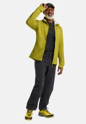 Veste en polaire vert citron, chemise noire, pantalon gris et chaussures de randonnée jaunes. Avec une fermeture éclair sur le devant et des poches, et une capuche.