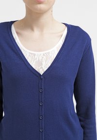 Námornícky modrý kardigán z úpletovej tkaniny s výstrihom do V, dlhými rukávmi a šiestimi gombíkmi vpredu, vrstvený cez čipkový top.
