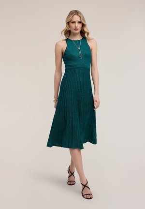 Frau trägt ärmelloses türkisfarbenes Midikleid mit plissiertem Rock und schwarzen Riemchenabsätzen, dazu goldener Schmuck, geht nach vorne.