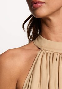 Beige halter dress with pleated texture, featuring a round neckline and a smooth fabric finish. Exposed shoulder and neck area visible.