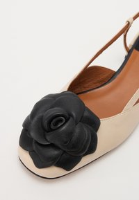 Beige slingback shoe with a glossy black leather rose detail at the toe. Features a flat sole and a smooth, tan interior lining.