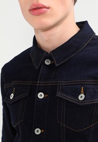 Ung person som bär en mörkblå denimjacka med kontrasterande orange sömmar och silverknappar, visad från halsen till mitten av torse.