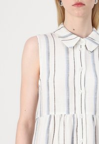 Femme portant un chemisier blanc sans manches avec des rayures verticales bleues et noires et un col pointu, boutonné devant.