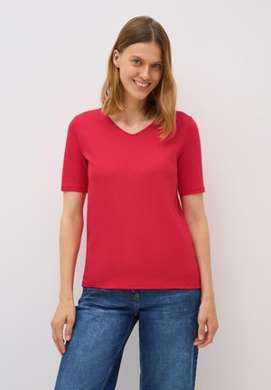 Rød kortærmet T-shirt med V-hals, lavet af glat stof. Båret med blå jeans, der fremhæver en tætsiddende silhuet.