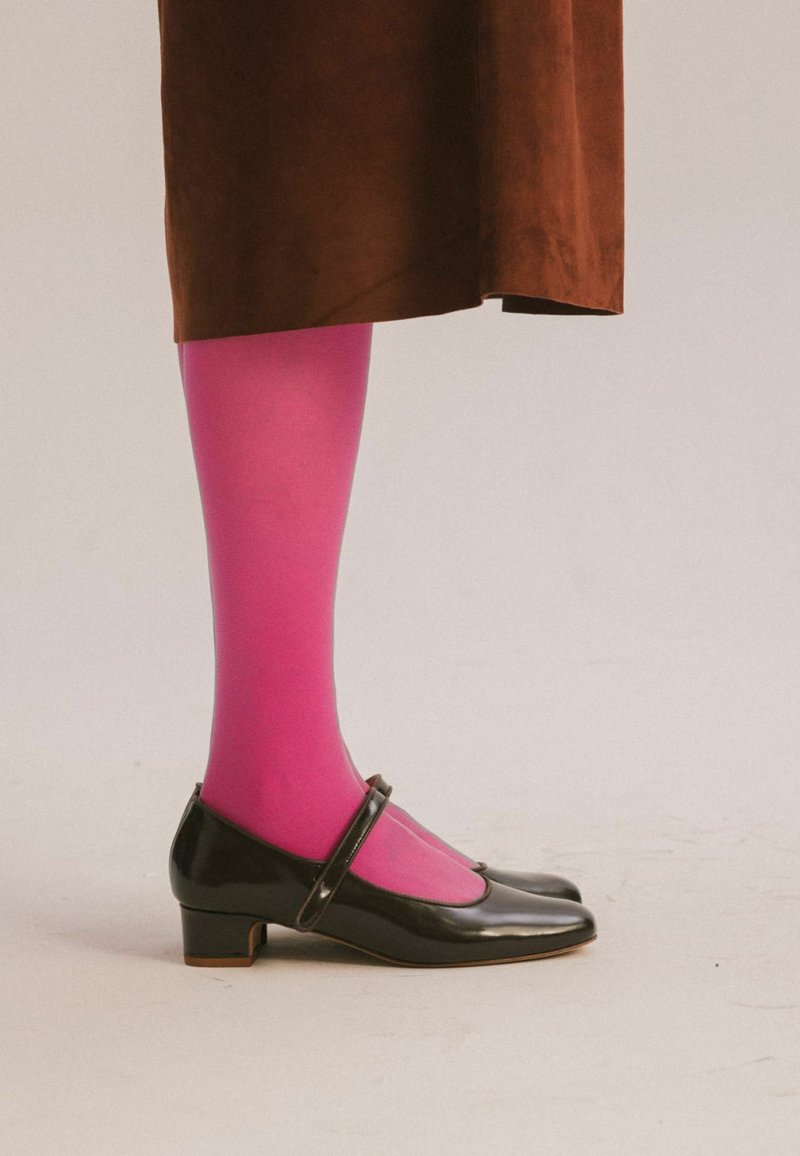 Gamba in collant rosa acceso che indossa una gonna marrone e scarpe nere Mary Jane con tacco basso su sfondo chiaro uniforme.