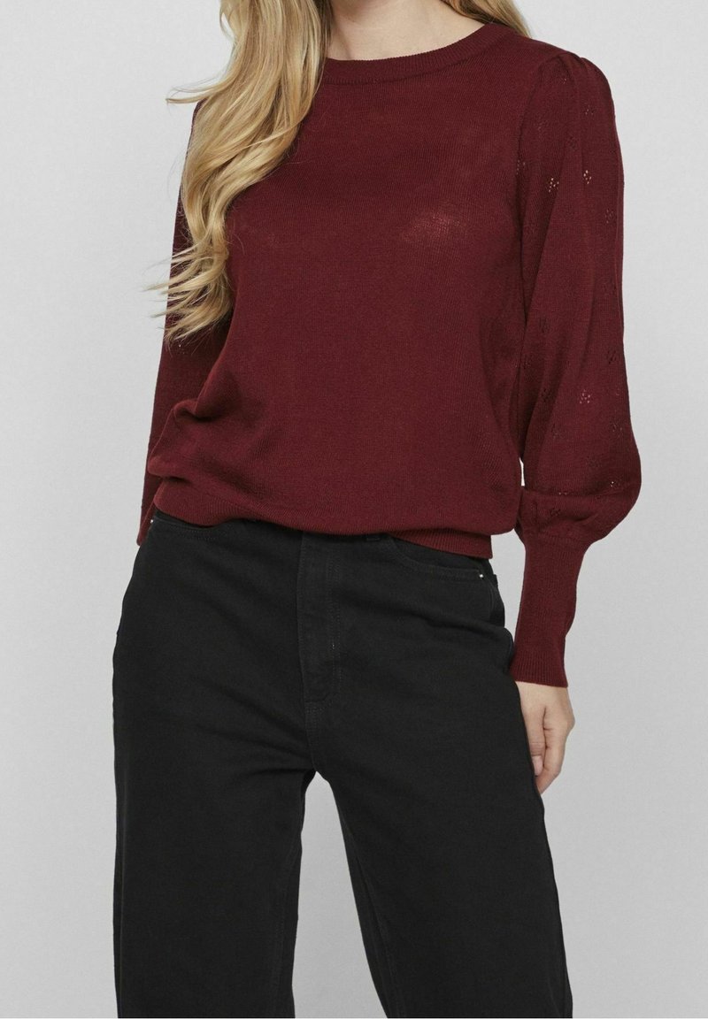Femme portant un pull en maille bordeaux avec des manches bouffantes et un pantalon noir taille haute sur un fond clair uni.