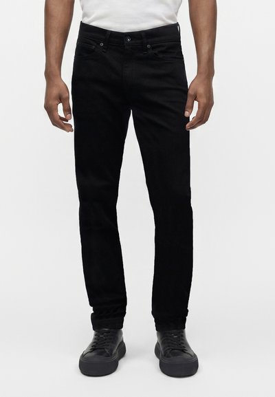 Homme portant un jean noir slim, des baskets noires et une chemise blanche sur un fond clair uni.