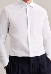Camicia bianca a maniche lunghe con colletto appuntito e tessuto liscio; indossata con pantaloni scuri.