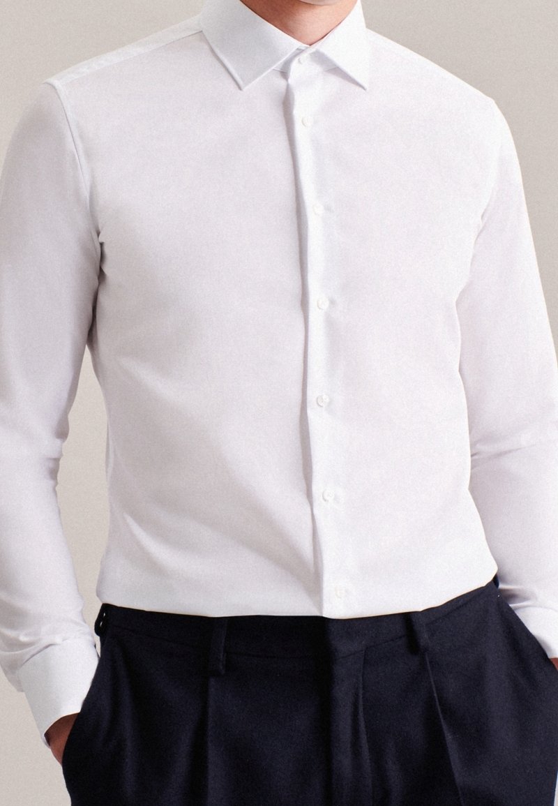 Camicia bianca a maniche lunghe con colletto appuntito e tessuto liscio; indossata con pantaloni scuri.