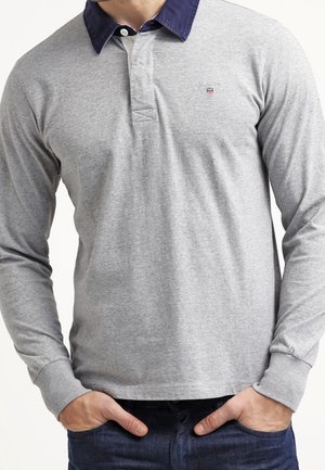Mand iført en lysegrå langærmet poloshirt med mørkeblå krave, hænderne i lommerne, synlig fra hals til hofte.