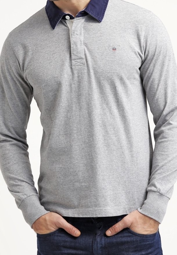 Homme portant un polo à manches longues gris clair avec un col bleu foncé, mains dans les poches, visible du cou aux hanches.