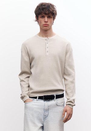 Junger Mann mit lockigem Haar, der ein beiges Langarm-Henley-Shirt, hellblaue Jeans und schwarzen Gürtel trägt, mit einer Hand in der Tasche stehend.