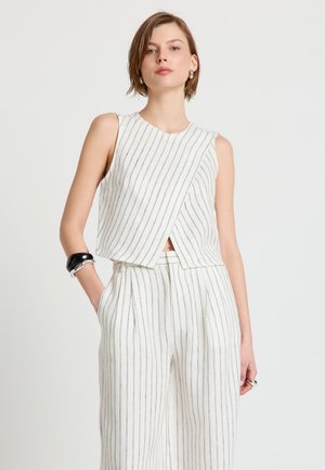 Frau trägt ein weißes ärmelloses Oberteil und weite Hosen mit dünnen vertikalen schwarzen Streifen, steht mit einer Hand in der Tasche.