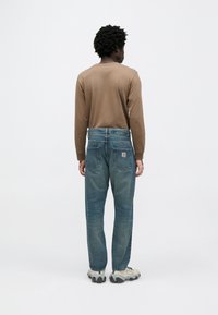 Brązowa koszula z długim rękawem w połączeniu z niebieskimi, wysokim stanem jeansami. Jeansy mają widoczne przeszycia, tylne kieszenie i naszywkę z logo marki. Całość dopełniają trampki.