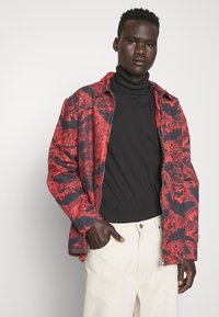 Giacca a motivo rosso e nero, in tessuto leggero, vestibilità rilassata, colletto aperto, abbinata a un dolcevita nero e a pantaloni color crema.