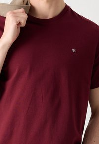 Burgundské tričko vyrobené z bavlny, s kulatým výstřihem, krátkými rukávy a malým vyšitým logem na levém prsu. Hladká textura.