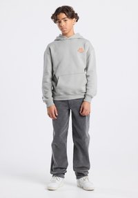 Grijze hoodie met een buidelzak en een oranje logo, gecombineerd met grijze broek en witte sneakers, allemaal tegen een effen achtergrond.