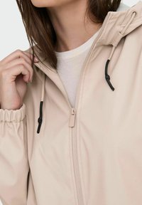 Chaqueta de color beige claro con cremallera y capucha. Cuenta con puños elásticos, ajuste con cordón y una textura de tela suave.