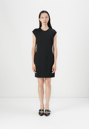 Abito nero a maniche corte con scollatura rotonda, silhouette aderente e tessuto liscio. Abbinato a scarpe nere con punta affusolata e cinturini alla caviglia.