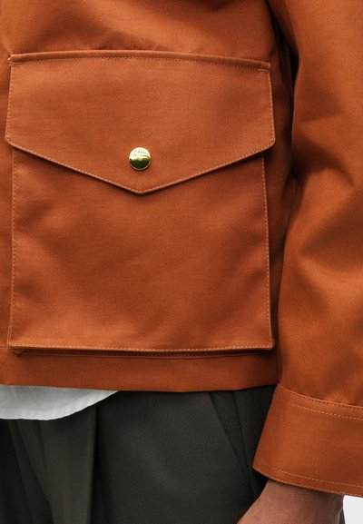 Barna dzseki, nagy elülső zsebbel, lekerekített fedele és arany színű patenttal. Sima anyagtextúra és szabott ujjak.