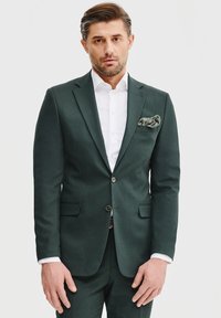 Ciemnozielony garnitur o slim fit, z zapięciem na dwa guziki, klapami typu notch i białą koszulą. Dodatkowa chusteczka w kieszeni zdobiona jest wzorem kwiatowym.