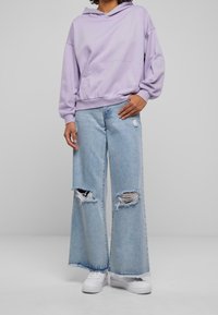 Sudadera con capucha de color lavanda combinada con jeans de pierna ancha desgastados de color azul claro. Zapatillas blancas completan el conjunto.