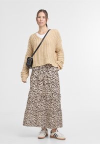 Woman standing wearing beige chunky knit sweater, white shirt, leopard print long skirt, sneakers, and black crossbody bag.