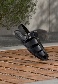 Μαύρα δερμάτινα σανδάλια με πλεκτό σχέδιο, ανοιχτό δάχτυλο και πλαϊνή αγκράφα, τοποθετημένα πάνω σε ξύλινες πλακώνες με μικρές πέτρες.