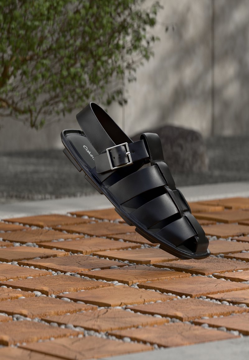 Μαύρα δερμάτινα σανδάλια με πλεκτό σχέδιο, ανοιχτό δάχτυλο και πλαϊνή αγκράφα, τοποθετημένα πάνω σε ξύλινες πλακ ώνες με μικρές πέτρες.