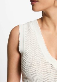 Top bianco lavorato a maglia con motivo a zigzag, scollatura a V profonda e orli ondulati sulle maniche. Offre un aspetto leggero e testurizzato.