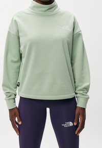 Sudadera verde de cuello alto con mangas largas, que presenta un pequeño logo blanco en el pecho. Combinada con leggins oscuros de color púrpura, también con logo.