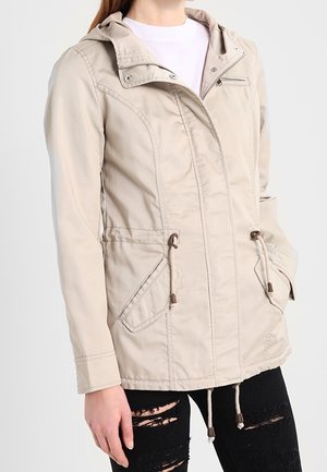 Femme portant une veste à capuche beige avec fermeture éclair et poches à cordon, associée à un jean noir déchiré, debout devant un fond uni.