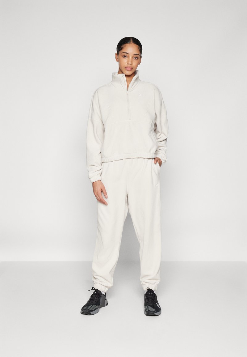 Jasno beżowy polarowy sweter z krótkim zamkiem błyskawicznym i prążkowanym dołem, w komplecie z dopasowanymi joggerami z mankietami. Czarne buty sportowe dopełniają zestaw.