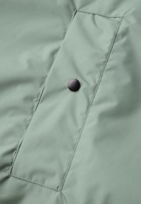 Padded jacket w jasnozielonym materiale o gładkiej fakturze, z boczną kieszenią i okrągłym czarnym zatrzaskiem.