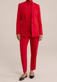 Röd skräddarsydd kostym med en blazer med en knapp, matchande figursydda byxor och en polotröja. Platta svarta skor fulländar outfiten.